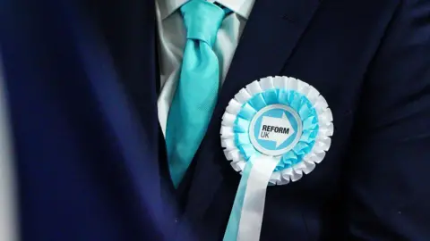 A blue and white Reform UK rosette is pinned to a dark blue suit with a light blue tie.