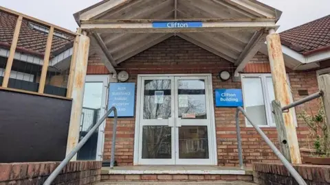 The porch of a brick building with blue NHS-style signage. It has metal hand rails and a glass door framed in white. One of the signs reads "The Clifton Building" . 