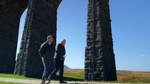 Nick Wooley/BBC Correspondent Danny Savage and Peter Myers walk below the viaduct, in the background are green rolling fields and a clear blue sky