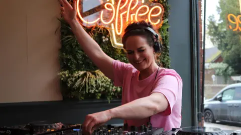 A close up of Lauren playing a set at a coffee shop in St Albans. She is holding up her arm and twiddling a knob and looks like she is enjoying herself. 