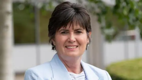 Solihull Council A woman with short black hair, a blue jacket, silver necklace and white top, smiles as she stands outside.
