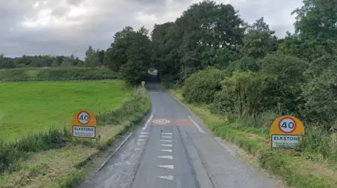 A country lane with a field on the left side and trees and bushes to the right. There are signs that read "Elkstone, please drive carefully through the village" with 40 miles per hour signs above. The lane ascends up a sloping hill, with trees covering the top end of the lane.