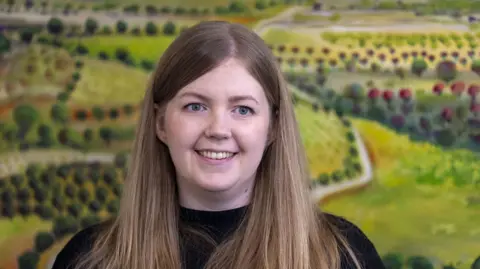 A woman with long brown hair and wearing a dark top smiled at the camera. A backdrop shows a rural scene