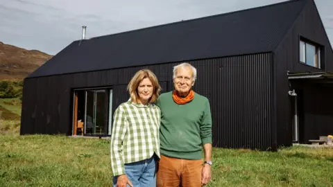 IWC Media Two people stand side by side in a grassy outdoor area in front of a modern, dark-clad house with a sloped roof.
The building features large windows, a metal chimney pipe, and a minimalist design set against a rural hillside landscape.
The scene is lit by natural daylight, with muted skies and earthy tones highlighting the contrast between the black structure and the surrounding greenery.