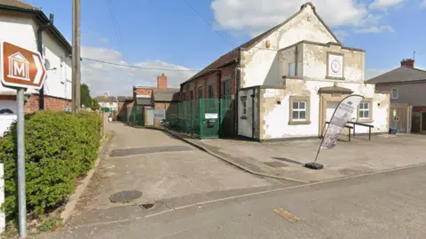 Google Streetview image of the old village hall - a single storey brick building with a weatherbeaten whitewashed entrance. Two signs point down a side road to the mining museum.