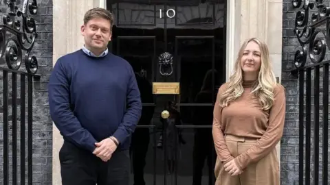 The Menai-Davis family Ceri and Frances Menai-Davis stand in front of the door of 10 Downing Street. Ceri is wearing a blue jumper and black trousers while Frances is wearing a brown top and brown trousers.