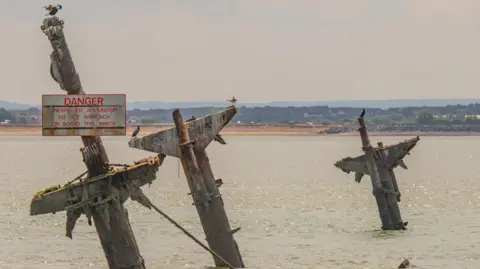 Three weathered masts from the SS Richard Montgomery rise from the water in the Thames Estuary. A warning sign attached to one mast reads: "Danger Unexploded Ammunition Do Not Approach Or Board This Wreck". Several seabirds are perched on the structures, with shoreline and buildings visible in the distance, in the photograph from 1 Aug 2022.