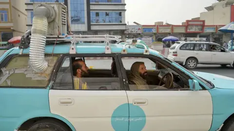 A light-blue Afghan taxi is with a hand-made air-cooler system fixed on top of it, - with its exhaust tube taped onto the window - passing through a market in Kandahar on 8 July. The driver is seen ferrying a male passenger. 