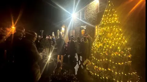 A group of people stand around a decorated Christmas tree outside, illuminated by lights and a single street lamp.