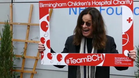 Birmingham Children's Hospital and Charity Ozzy Osbourne holding up in front of him a piece of cardboard, where most of the middle has been cut out, allowing us to see his face. It includes the words Birmingham Children's Hospital Charity.   