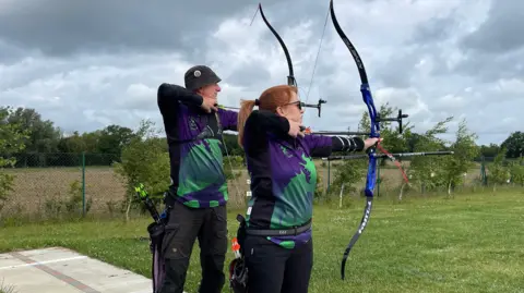 Edd Smith/BBC Two people on the shooting line at an archery club with bows and arrows who are both stood sideways ready to hit a target.