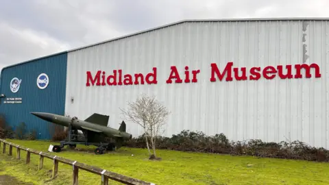 A large white metal hangar with the words Midland Air Museum in red on the side and a green military missile on grass outside