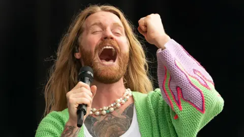 PA Media Sam Ryder performing on stage. He is singing with his eyes closed, his mouth wide and is holding his fist in the air. He has very long fair hair, a beard and is wearing a pearl necklace and green knitted cardigan with purple flames on the end of the sleeves and has a tattooed chest