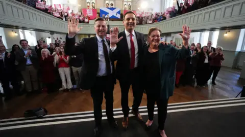 PA Media Scottish Labour leader Anas Sarwar, accompanied by Scottish Secretary Douglas Alexander (left) and deputy leader of the Scottish Labour Party, Dame Jackie Baillie, during an eve of poll rally at Adelaide Place Baptist Church in Glasgow, on the last day of campaigning ahead of the Holyrood elections on Thursday. They are in dark suits and are waving with a crowd behind them.