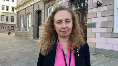 BBC Deputy Inna Gardiner is wearing a black blazer and a pink top. She is standing in Jersey's Royal Square.