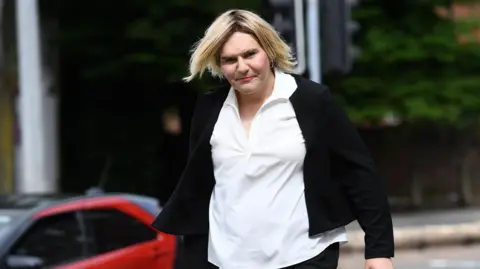 A picture of a transgender woman with short straight blonde hair, wearing make up, hoop earrings, a white blouse and a black cardigan. Katie Wallis is walking into court and looking at the camera.
