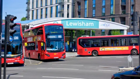Three buses drive past the entrance to Lewisham DLR station on a sunny day. There are residential blocks in the background. 
