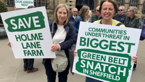 Two women standing with a group of campaigners outside Sheffield Town Hall. They are holding large green and white placards which call for the green belt to be protected: One reads: Save Holme Lane Farm, and the other reads: Communities under threat in biggest green belt snatch in Sheffield.