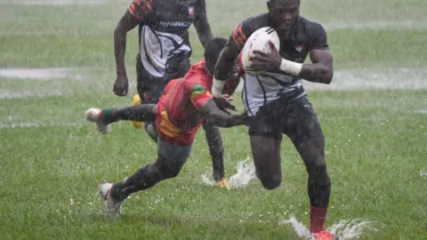 AFP A man running with a rugby ball. It is wet and there is rain splashing. A player behind him is diving, trying to stop his from running.