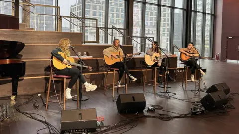 Kezia Gill, Jess Thristan, Demi Mariner, and Jade Helliwell (left to right) playing the guitar and singing in preparation for a gig at Birmingham Symphony Hall. The singers are all pictured sitting down on stools holding their instruments with microphones on stands in front of them. To the left of the photo is a grand piano and the buildings of Birmingham city centre can be seen out of a large window on the right