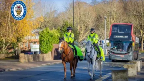 Tow mounted police officers on a brown horse and another is riding a white horse. They are riding in front of a National express West Midlands red and black bus