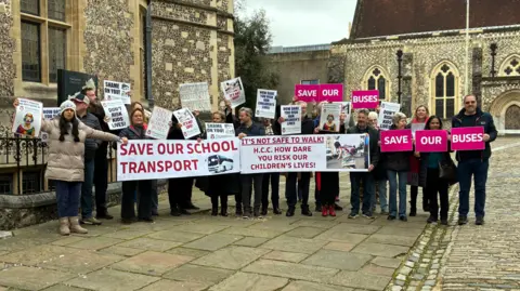 A group of protesters outside Winchester Registry office, holding white and pink cards which read "save our buses", "save our school transport" and "It's not safe to walk, HCC how dare you risk our children's lives"