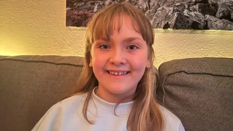 Emberly, a girl with blond hair and a white sweatshirt sat on a sofa smiling towards the camera.