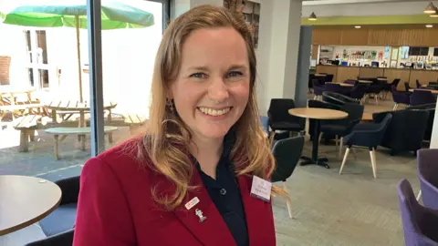 A woman in a red blazer jacket and blue polo shirt smiles at the camera. She is shoulder length hair. She is in a coffee shop surrounded by tables and chairs.