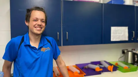 Exploris seal worker Aran Pollock smiling at camera. He is wearing a blue pop shirt. He has brown hair. There is seal feeding equipment in the background sitting on orange, purple and a green tray.