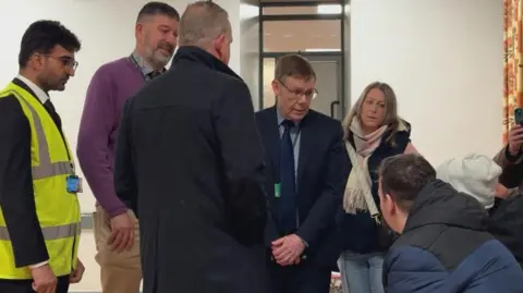 A group of men and women standing around, negotiating inside a community centre which was hosted a council meeting.