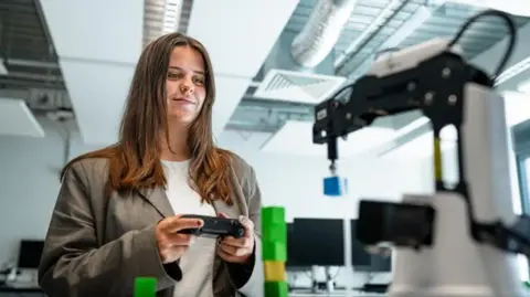 Harper Adams University A woman with long brown hair, a white top and brown jacket holding a small black device in a white room with mechanical devices in it