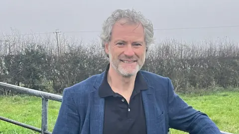 A man standing in front of an iron gate in a field, with a hedge row behind him. He is wearing a navy blue suit and a black shirt. 