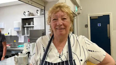 A woman with short blonde hair is wearing a white and black shirt and an apron. She is standing in front of a kitchen.