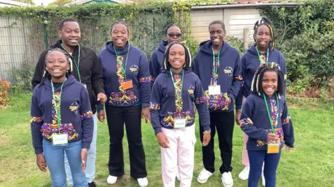 Luke Deal/BBC Members of the choir stand outside and are all smiling at the camera. With the exception of one man, they are all wearing navy hoodies with a colourful pattern down the middle. They are also wearing green lanyards. The outdoor setting they are in has grass on the floor and a fence behind them.