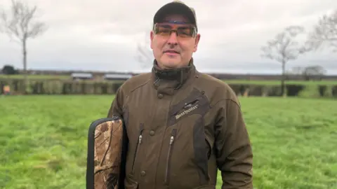 Picture of Gary McCartney, Director of Countryside Alliance Ireland. He is wearing a khaki jacket and cap, with protective eye wear on.
He is carrying a rifle bag.