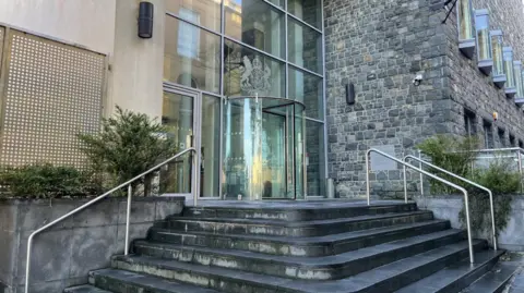 BBC Frontage of Guernsey royal courts. Steps leading up to a glass entrance with a crest above it.