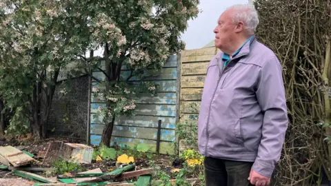 Mr Campbell is wearing a mauve coat and dark trousers. He looks to the side as he views his allotment. There is debris including wooden fence panels on the ground.