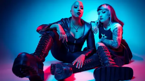 Paul Harries Sophie is wearing an all black leather outfit, with knee high lace up boots and a zip up leather top. Her head is shaved and she is wearing rings on most of her fingers, a necklace and dark lipstick. Yasmine has shoulder length blond and black hair with a short fringe. They have a sleeve of tattoos, a black and white t-shirt, black skirt and knee high black buckle up boots. They are both sat on the floor. 