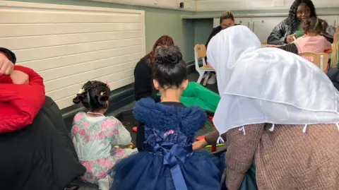 Shariqua Ahmed/BBC A woman in a white scarf can be seen with two girls playing on a black board on the floor. Their backs are facing the camera. 