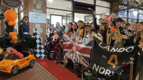 The Lord Lieutenant of Somerset smiles next to a road sign that says Lando Lane while a crowd of young people wave chequered flags and hold Lando Norris flags