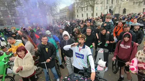 A man with DJ decks and a cat on a bicycle holds a microphone as he addresses a crowd of people with bikes in London