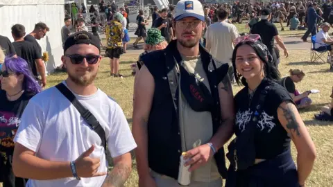 Friends Jordan, Alistair and Chloe stand in a line with the Download Festival in the background, with a crowd behind them. Jordan wears a reversed baseball cap, sunglasses and a white t-shirt. He's got a bag strap across his body and he's holding his right hand in front of his torso - pulling the metal sign with his ring and pinky fingers extended. Alistair's taller than same and wearing a forwards trucker cap and black denim waistcoat. Chloe's wearing mainly black with pink sunglasses perched on her head as she smiles.