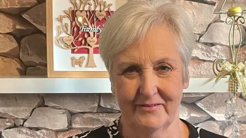 Wendy Falconer standing in a living room. She is middle-aged, with short blonde hair.