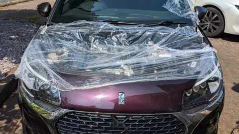 A car which was stripped of its bonnet and is now covered in cling film, waiting to be repaired