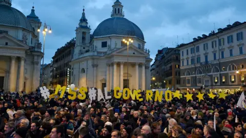 Getty Images A large crowd gathers in a city square. Among them are individual letters arranged to say : "M5S al governo *****"