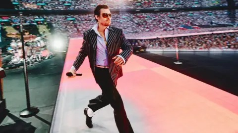 Harry Styles strides onto the stage at Wembley Stadium, wearing a black and white pinstripe jacket and a wide-collared blue shirt. Spotlights illuminate the audience, and the singer is wearing aviator sunglasses.