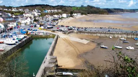 Charles Hawes/Getty Images This image shows Saundersfoot beach, the sand is beige and there's little boats docked. The sky is blue with some clouds and there's greenery in the distance. There's a stone wall separating the sandy beach from a collection of brightly coloured buildings. 