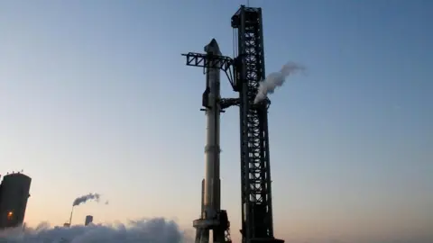 Roket Starship Reuters SpaceX berdiri vertikal di landasan peluncuran, dengan latar belakang langit biru cerah