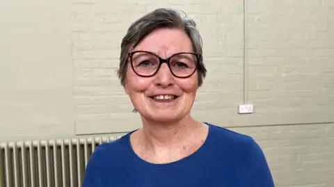 Ellen Knight/BBC Photo of Morris looking into the camera and smiling. She is wearing a dark blue, long sleeve top and a pair of dark-framed classes. Morris has short, light brown hair. She is photographed in front of a brick wall that's been painted light green. 
