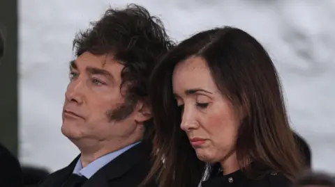 Reuters The president of Argentina, Javier Milei, and Vice-President Victoria Villarruel attend a commemorative ceremony in Buenos Aires on 18 March 2024.
They are standing next to each other. Villarruel is looking down and Milei is looking straight ahead. Both are wearing black jackets.